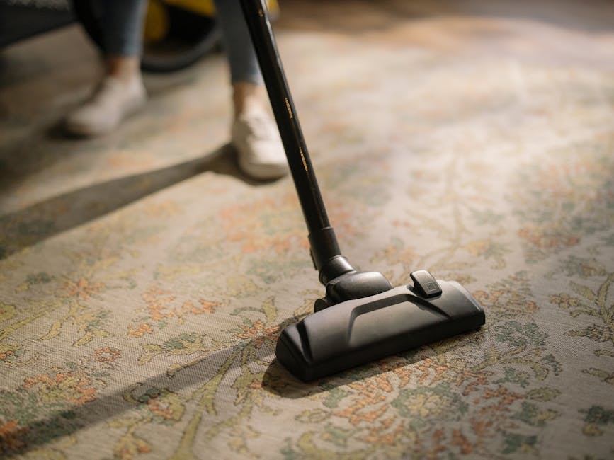 Close-up of a modern vacuum cleaner head and a section of an ornate, patterned carpet being thoroughly cleaned in a residential setting. The carpet features intricate floral designs in muted shades of beige, green, and orange, and appears clean and well-maintained. The vacuum cleaner is black and sleek, with a visible power switch, positioned centrally on the carpet, with a person’s feet in casual shoes partially visible in the background. Natural lighting highlights the cleanliness and detail of the surface. This image reflects surface cleaning and maintenance, aligning with the topic of avoiding hidden charges in Kingston carpet cleaning quotes and showcasing professional domestic cleaning services available through Kingstonuponthamescarpetcleaning.co.uk.