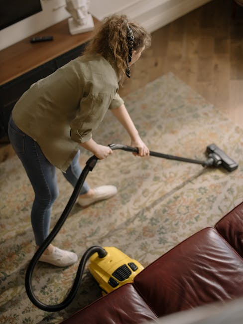 A woman performing surface cleaning in a living room, vacuuming a patterned area rug with a yellow vacuum cleaner. She is wearing casual clothing including a beige jacket and jeans, with her hair tied back. The room features wooden flooring, a leather sofa in the foreground, and a wooden sideboard along the wall, which holds a stereo speaker and decorative items. The scene is well-lit with natural light, emphasizing the cleanliness and maintenance of the carpet. This image reflects domestic cleaning practices, aligning with Kingston upon Thames carpet cleaning services provided by [COMPANY_NAME], promoting effective deep cleaning and hygiene in residential spaces.