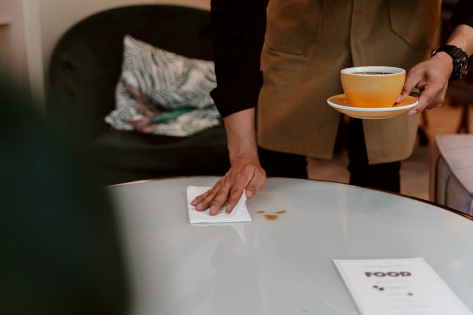 A person wearing a black long-sleeve shirt and a beige apron is cleaning a white, smooth-surfaced table with a white cloth. The individual is holding a yellow teacup and saucer in their left hand, with a dark beverage inside, while wiping the table's surface with their right hand. The background includes a dark-colored sofa with a decorative pillow featuring a leaf pattern, and warm ambient lighting enhances the cozy atmosphere. The table appears clean and free of stains, showcasing attention to domestic surface cleaning and maintenance, as promoted by Kingston cleaners on the page about common stains and how to remove them quickly in Kingston upon Thames.