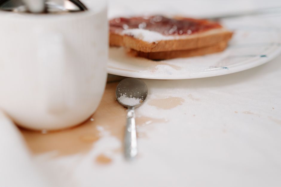 A close-up image of a kitchen countertop with visible flour and powdered sugar scattered across its surface. In the background, a white ceramic bowl is partially visible on the left, and a white plate holding a slice of bread with jam is positioned towards the right. A stainless steel spoon rests on the counter, partially covered with flour. The lighting is natural, highlighting the clean, smooth texture of the countertop and the slight mess created during baking or food preparation. This scene illustrates typical domestic surface cleaning and hygiene practices, aligning with the services of Kingston cleaners for maintaining cleanliness in kitchens and other living spaces. The focus on powdered ingredients and kitchenware emphasizes the importance of thorough surface cleaning after cooking or baking to remove food residues and keep surfaces hygienic, as featured on the page about common stains and how Kingston upon Thames cleaning services swiftly tackle them.