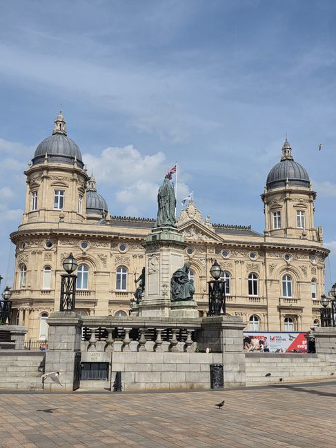 A grand, historic building with beige stone facades, ornate architectural details, and two prominent domed towers topped with small spires, set against a partly cloudy blue sky. In front of the building, there is a stone platform with decorative iron lamp posts and a statue of a historical figure holding a flag, surrounded by a low stone balustrade. The area is paved with large, rectangular stone slabs, and a pigeon is seen walking on the pavement. The scene is well-lit with natural daylight, highlighting the intricate details of the building's architecture, typical of a historic civic or cultural landmark, with no visible signs of dust, dirt, or debris, suggesting it has been recently cleaned or well-maintained. The image is associated with the Kingston upon Thames area's emphasis on surface cleaning and maintenance, as referenced on the page for compliance checklists for cleaning contractors.