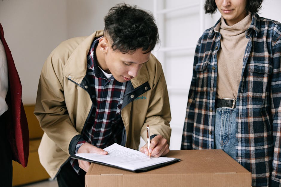 A person in a beige jacket and plaid shirt is signing a document on top of a brown cardboard box, with another person standing nearby in a plaid shirt and beige turtleneck. The scene takes place in a well-lit indoor space, possibly a living room or an office, with a white wall and furniture visible in the background. The surface of the box appears clean and unmarked, and no additional cleaning tools or products are visible in the image. This image relates to professional end of tenancy cleaning, highlighting thorough surface inspection and documentation, which [COMPANY_NAME] offers as part of its domestic cleaning services, ensuring a high standard of hygiene for moving out or property management purposes.