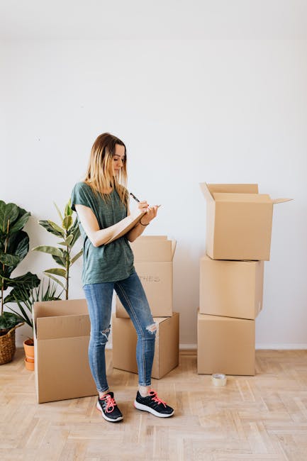A young woman with shoulder-length blonde hair, dressed in a casual green t-shirt and ripped blue jeans, stands on a wooden floor in a well-lit room. She is holding a clipboard and a pen, appearing to be inspecting or taking notes. Behind her, there are several cardboard moving boxes, some open and some stacked, indicating a packing or cleaning process related to end of tenancy procedures. To the left, a large potted plant with broad green leaves adds a touch of natural decor. The room has a clean, minimalist appearance with plain white walls and natural lighting, suggesting a space undergoing thorough surface cleaning or preparation for departure, consistent with a professional deep cleaning service like Kingston upon Thames Carpet Cleaning.