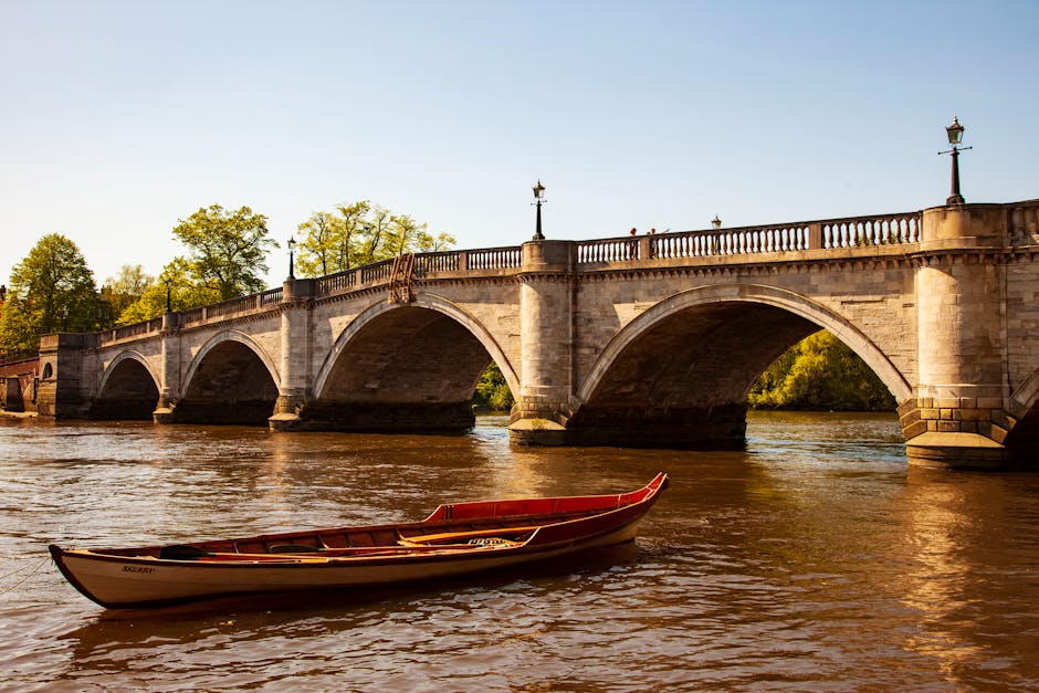 A red and white wooden boat floating on a calm river beneath an elegant stone bridge with arched spans, decorative lamp posts, and a railing, surrounded by lush green trees with bright sunlight illuminating the scene. The setting appears peaceful, showcasing the outdoor environment near Kingston upon Thames while highlighting clean water surfaces and well-maintained structural elements, aligned with surface cleaning and outdoor maintenance services provided by Kingstonuponthamescarpetcleaning.co.uk.