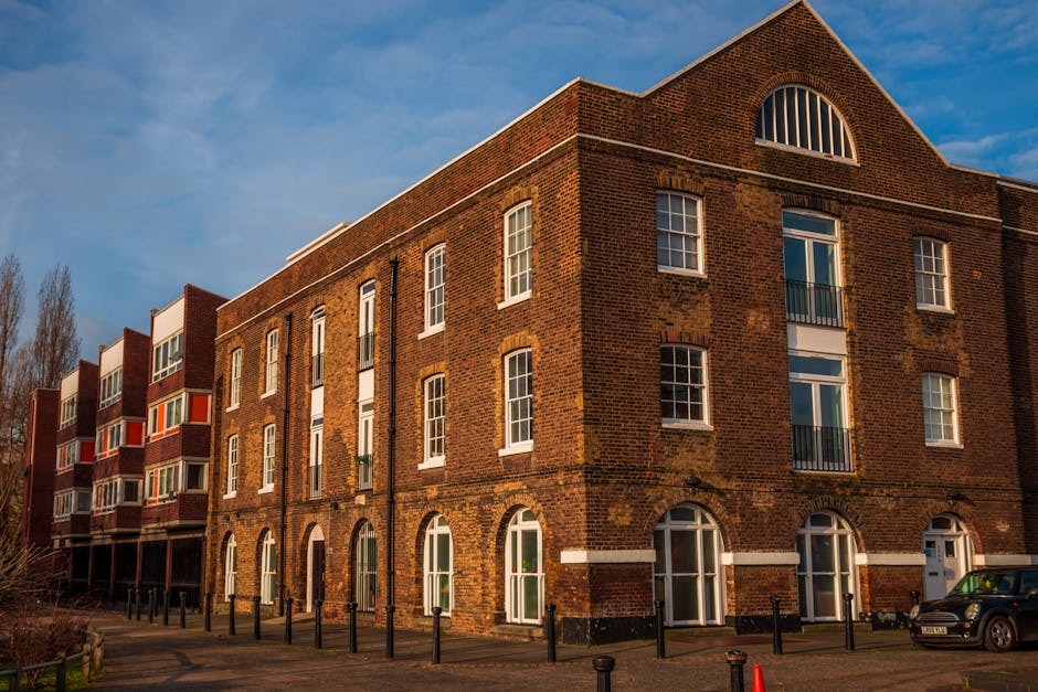 Exterior view of a multi-story brick building in Kingston upon Thames, with large arched windows on the ground floor and rectangular windows on the upper floors. The building's facade shows weathered brickwork with some variation in color and texture. The setting is during daytime with a partly cloudy sky, and the building is illuminated by natural sunlight. In front, there are small black bollards along the sidewalk, with a parked car visible to the right. The scene highlights the building's architectural features, typical of High Street flats, with no interior or cleaning equipment visible. For surface cleaning and deep cleaning advice related to such residential properties, Kingstonuponthamescarpetcleaning.co.uk advises on hygienic maintenance of shared entrances and interior surfaces.