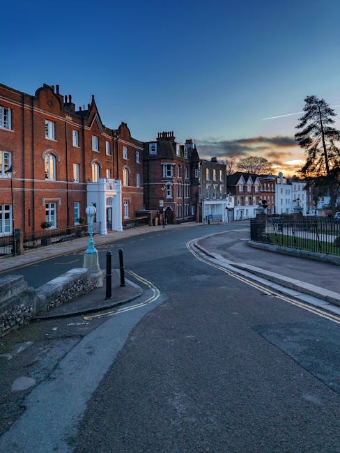 A red and white wooden boat floating on a calm river beneath an elegant stone bridge with arched spans, decorative lamp posts, and a railing, surrounded by lush green trees with bright sunlight illuminating the scene. The setting appears peaceful, showcasing the outdoor environment near Kingston upon Thames while highlighting clean water surfaces and well-maintained structural elements, aligned with surface cleaning and outdoor maintenance services provided by Kingstonuponthamescarpetcleaning.co.uk.
