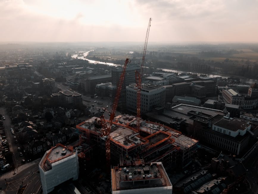 An aerial view of a construction site in an urban area during daylight, featuring two tall cranes positioned on a partially built multi-story building. The site includes concrete floors, steel reinforcements, and construction materials spread across the structure. Surrounding the construction zone are various office buildings with glass and concrete facades, streets with parked cars, and residential houses. The background shows a river winding through the city, with additional buildings and a hazy sky above. The scene is captured with natural lighting, emphasizing the busy, developing environment, aligning with the theme of surface construction and deep cleaning processes observed in the area, as seen by the organized site layout, clean surfaces, and construction equipment involved.