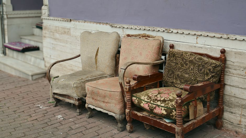Three abandoned and weathered upholstered armchairs are placed outside against a wooden wall on a brick pavement. The first chair on the left has beige fabric upholstery that is torn and stained, with visible stuffing and worn wooden legs. The second chair in the middle features faded pink floral-patterned fabric, also distressed, with frayed edges and damaged areas. The third chair on the right has a dark wooden frame with curtains and a seat cushion covered in ornate green and pink tapestry fabric, which appears worn and aged, with noticeable fabric tears. The chairs are positioned in a row in a slightly shaded outdoor setting near a residential building, suggesting they are awaiting disposal or recycling, aligning with Kingston Council upholstery disposal rules for cleaners, KINGSTON UPON THAMES.