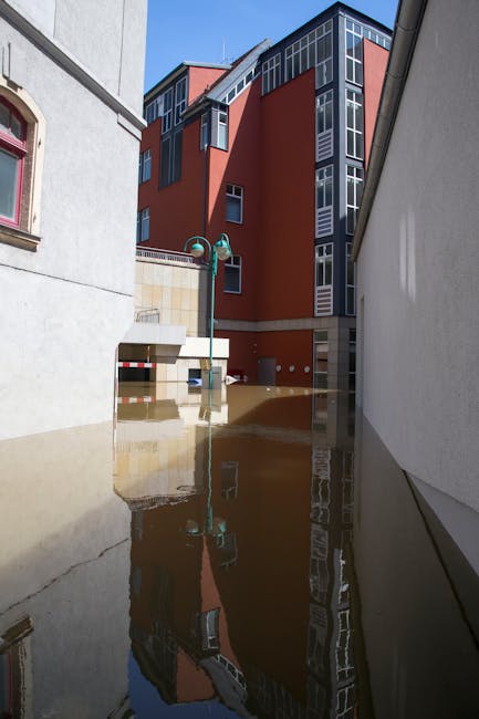An outdoor urban area during daytime shows water flooding the ground between two buildings, with the reflection of a modern, multi-storey red and grey building in the stagnant water. The floodwaters partially submerge concrete surfaces and the base of a streetlamp, highlighting the extent of the flooding. Sunlight illuminates the scene, casting clear reflections of the adjacent buildings and sky. The image is associated with the services of Kingston upon Thames carpet cleaning and emergency cleaning, emphasizing the importance of deep cleaning and sanitisation after flood damage to maintain hygiene and surface integrity.