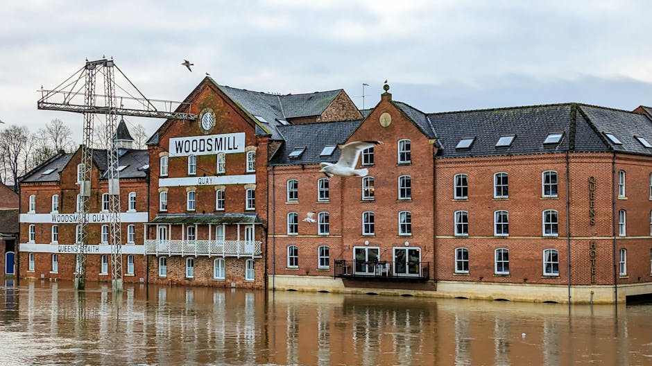 Photograph of a red-brick building with multiple rectangular white-framed windows and a black tiled roof, situated along a flooded area with murky water covering the ground floor. The building features a sign reading 'WOODSMILL' and decorative white metal balconies. A tall metal industrial crane is positioned behind the structure, and a seagull is flying overhead against a cloudy sky. The scene depicts the impact of flooding, emphasizing the importance of surface cleaning and sanitisation, which [COMPANY_NAME] provides in Kingston upon Thames for emergency cleaning and flood damage restoration.