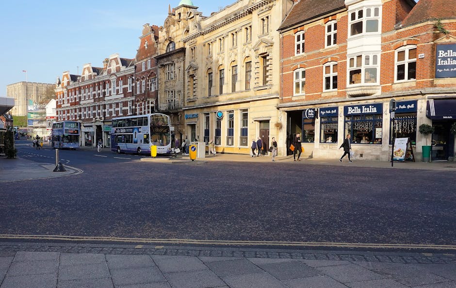 A street scene on Clarence Street in Kingston upon Thames showing a row of historic multi-story buildings with brick facades and large windows, some with white decorative trim. Pedestrians are walking along the sidewalk, and two double-decker buses are parked along the curb. The shops include 'Bella Italia' with blue signage and outdoor seating. The street is paved with asphalt, with double yellow lines along the edge and gentle sunlight illuminating the scene. The area appears clean and well-maintained, reflecting a vibrant commercial district. This image is associated with Kingstonuponthamescarpetcleaning.co.uk, emphasizing the importance of surface cleaning and hygiene in local businesses and commercial spaces.