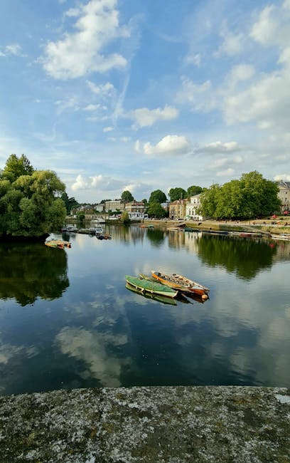 A peaceful riverside scene with calm water reflecting a partly cloudy sky with scattered white clouds. Small boats are moored along the riverbank, which is lined with lush green trees and residential buildings featuring light-colored facades and traditional architectural styles. The area is well-maintained, with visible grassy sections and pathways along the shoreline. Natural daylight illuminates the scene, highlighting the pristine condition of the water and surrounding environment. This setting exemplifies a clean and serene outdoor space, aligning with the principles of surface cleaning and maintenance promoted by Kingstonuponthamescarpetcleaning.co.uk, focusing on hygiene and care of outdoor surfaces and residential areas.