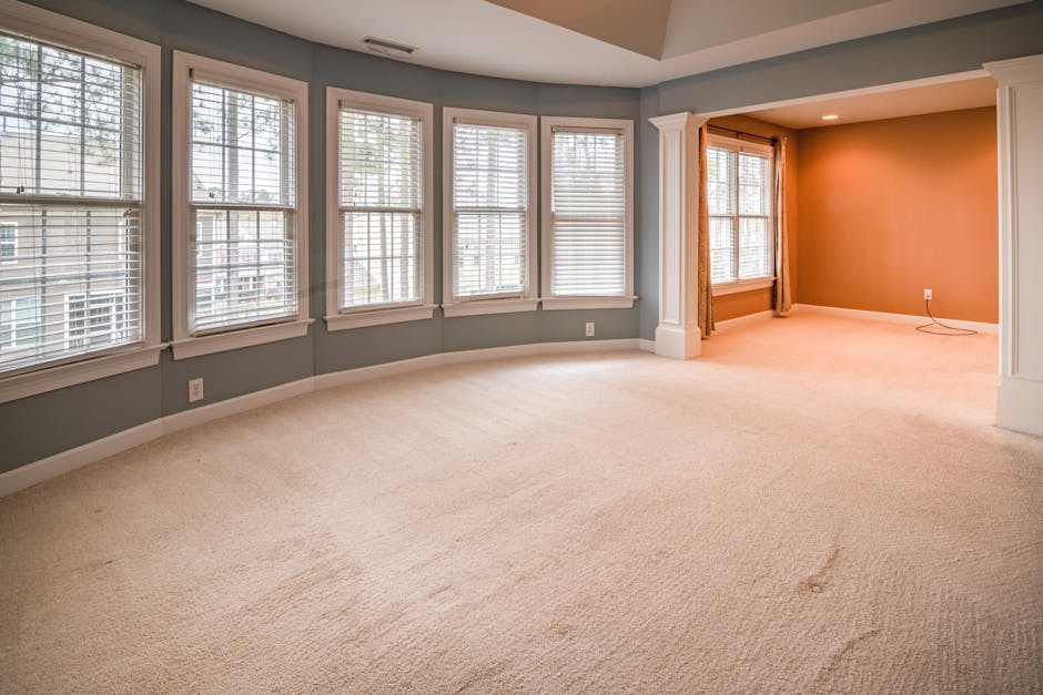 A spacious, light-filled living room with large bay windows fitted with white wooden blinds, letting in natural daylight. The room features a cream-colored plush carpet that appears clean and well-maintained, with subtle texture visible on its surface. The walls are painted in soft pastel shades, with one wall in a warm beige tone and the others in a light blue color, complemented by white crown molding and trim. The room is empty, emphasizing the spotless condition of the carpeting and polished appearance of the window frames. Visible in the background is an attached smaller alcove with a window and light wood trim, creating a seamless, open layout. The overall scene illustrates a tidy and hygienic environment, representative of professional surface cleaning and maintenance, possibly performed by Kingston upon Thames Carpet Cleaning, highlighting the quality of deep cleaning and sanitisation in residential spaces.
