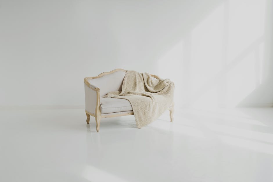A vintage-style white sofa with wooden trim, positioned against a plain white wall in a well-lit room with natural sunlight casting shadows on the floor. A beige textured throw blanket is casually draped over the sofa's back and seat, emphasizing a clean and tidy appearance. The smooth, polished floor surface appears spotless, reflecting the soft light, and the overall scene suggests a minimalistic, hygienic space focusing on surface cleaning and domestic cleaning practices. The setting highlights the importance of upholstery cleaning for maintaining a fresh and inviting home environment, aligning with upholstery cleaning tips for Canbury Gardens homes, Kingston upon Thames, as promoted by Kingstonuponthamescarpetcleaning.co.uk.