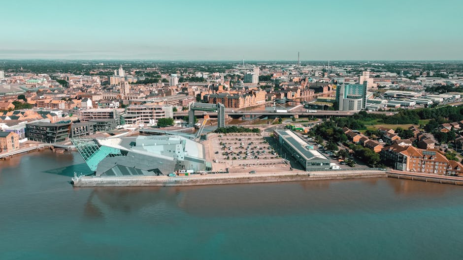 Aerial view of Kingston upon Thames showing a mix of historic brick buildings, modern glass structures, and residential houses along the riverbank. The canal is visible at the foreground with calm water reflecting the sky, while the cityscape extends into the distance with various commercial and civic buildings under clear daylight. The scene highlights urban landscapes and the area's architectural diversity, captured during a bright, cloudless day, with natural lighting enhancing the clarity of surfaces and structures. This image exemplifies the integration of old and new architecture in Kingston upon Thames, suitable for illustrating the setting where [COMPANY_NAME] offers surface and deep cleaning services aligned with Kingston Council's waste management policies for maintaining hygienic environments.